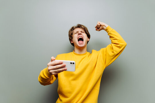 Emotional young man in yellow clothes rejoices in victory with a smartphone in his hands on a gray background. Mobile gaming. gamer won an online game on a smartphone.