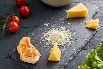 Ingredients for making salad. Piece and grated parmesan cheese on black serving board, cherry tomatoes, roasted chicken and lettuce leaves.