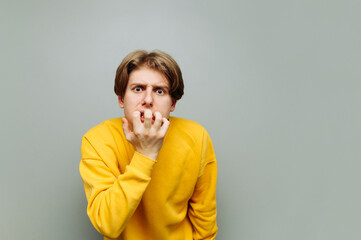 Portrait of a scared young man in a yellow sweatshirt on a gray background, looking at the camera and biting his fingers from nerves. Isolated.