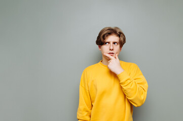 Pensive guy in casual clothes stands against a gray wall and looks away with reflections on copy space. Isolated.