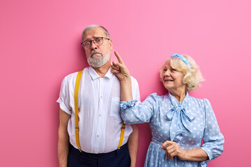 couple have fun,woman is playing with gray-haired man isolated on pink background, index finger at man's face
