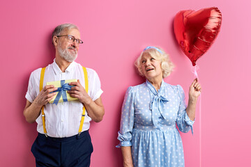 shy man stand with gift box for wife, attractive lady in dress holding air balloon, isolated on pink background. Valentine's Day concept