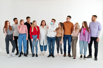 sick male in medical mask stand among healthy people looking at him. group of people stand in a row looking at male standing in center, isolated on white studio background