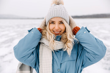 Portrait of a cheerful young blonde woman on cold winter day, smiling, adjusting hat.