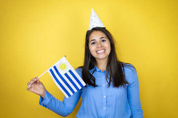Young caucasian woman wearing a birthday hat over isolated yellow background smiling and holding a Uruguay flag