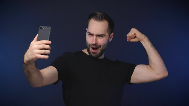 Funny bearded young man 20s in black casual t-shirt isolated on dark blue background studio. People lifestyle concept. Doing selfie shot on mobile phone showing thumb up victory sign biceps muscles