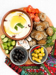 Roasted chickpeas falafel patties with a plate of Hummus, served with fresh vegetables and olives in a wooden plate over light background. Healthy vegan food, clean eating, dieting, top view