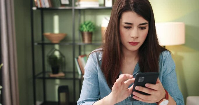 Close up of Caucasian beautiful young brunette woman sitting at desk and texting on cellphone using social network app surfing internet on smartphone in cozy cabinet, communication concept