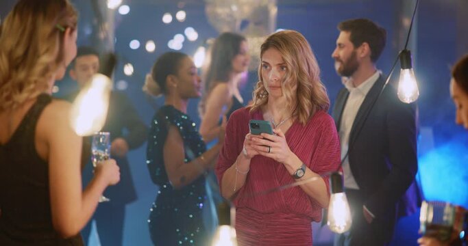 Beautiful Blonde Young Caucasian Woman Using Mobile Phone texting Messages in Crowd of Dancing People Celebrating Holiday. Corporate Party Concept.