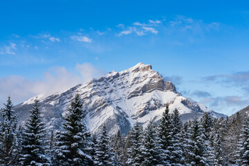Close up snow-covered Cascade Mountain with snowy forest over blue sky and white clouds in winter sunny day. Banff National Park beautiful landscape. Canadian Rockies, Alberta, Canada. - obrazy, fototapety, plakaty