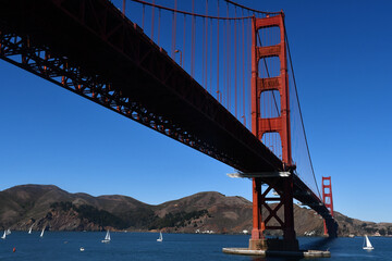 Golden Gate Bridge, seen from below, and white sailboats on the Golden Gate strait on a sunny day with a cloudless blue sky