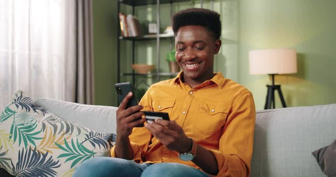 African American cheerful satisfied young handsome male resting sitting on sofa in room, browsing online on cellphone buying on internet making purchase using credit card. E-commerce, portrait concept