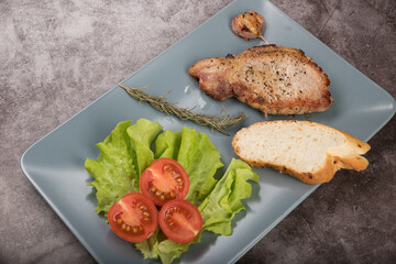 Roast pork loin, on a rectangular plate of gray color, with lettuce leaves, rosemary a piece of baguette and tomatoes, on a gray background