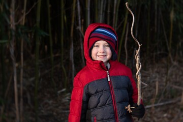 Young school age boy playing in the woods holding stick wearing winter jacket and hat