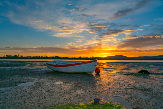 Backlighting of small boat at low tide on beach by sunrise across harbour and behind distant hills.