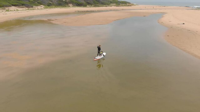 Aerial shot orbiting around a man in a wet suit on a stand up paddle board with his son down by the coastline.