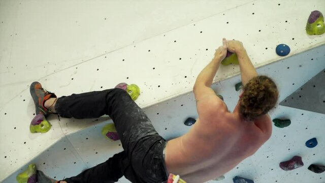 Tall athletic man indoor rock climbing with no ropes on over hung wall edge. Long curly hair in bobble in black pants with chalk bag on waist. 

1080P 25FPS 10 Bit SLOG2-REC709
