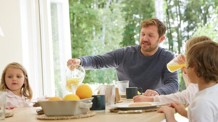 Father pouring orange juice as family wearing pyjamas sit around table enjoying pancake breakfast together - shot in slow motion