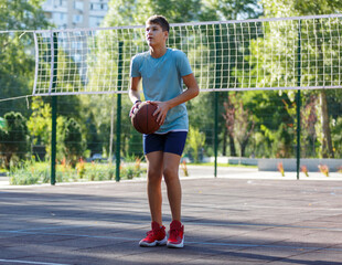 Cute young boy plays basketball on street playground in summer. Teenager in green t shirt with orange basketball ball outside. Hobby, active lifestyle, sport activity for kids.	