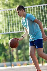 Fototapeta premium Cute young boy plays basketball on street playground in summer. Teenager in green t shirt with orange basketball ball outside. Hobby, active lifestyle, sport activity for kids. 