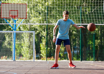 Obraz premium Cute young boy plays basketball on street playground in summer. Teenager in green t shirt with orange basketball ball outside. Hobby, active lifestyle, sport activity for kids. 