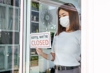 Woman with sad emotion wearing face mask turning a sign from open to closed through shop window. Small shop closed after covid-19 quarantine lockdown.