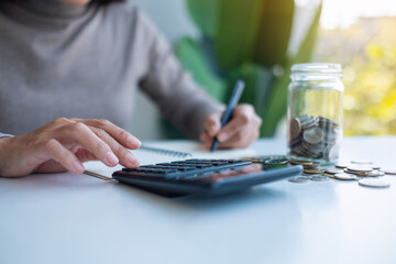 Closeup image of a woman saving money in a glass jar, calculating and taking note for financial concept