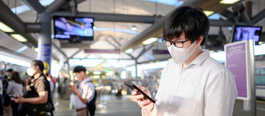 Asian man wearing surgical face mask using smartphone at skytrain station platform. Coronavirus (COVID-19) outbreak prevention in public transportation. Health awareness for pandemic protection