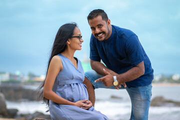 Young lovely married couple posing in a beach, pregnant with the first child, expecting a baby girl soon, parents to be soon, father pointing a finger to belly, outdoor Sri Lankan models photoshoot,