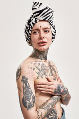 Tattoo and piercing. A white pierced and tattooed woman standing topless with a turban covering her breasts with hands wearing a denim overall looking into a camera with a mouth open slightly