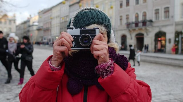 Portrait of old senior woman tourist taking pictures with photo camera, looking at camera using retro device in winter city center of Lviv, Ukraine. Photography, travelling, vacation. Active pensioner