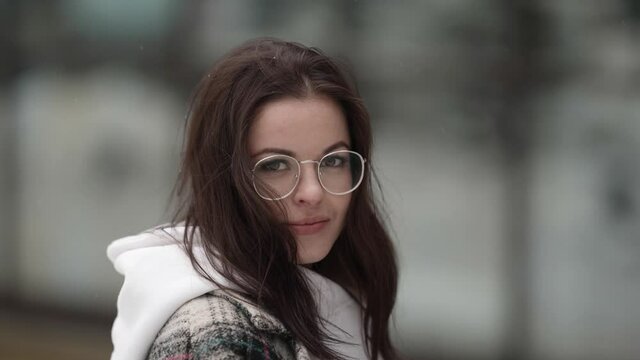 Portrait Of A Beautiful Young Dark-haired Woman Without A Hat, Wearing Round Glasses And A Plaid Coat, Walking On A Snowy Winter Day, Smiling And Winking.