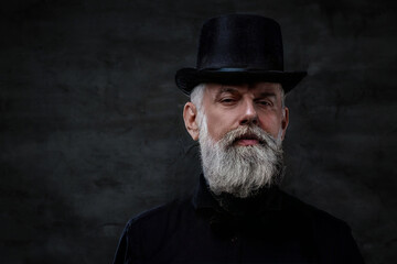 Dressed in old fashioned style clothing grandfather with top hat and gray hairs poses in dark background.