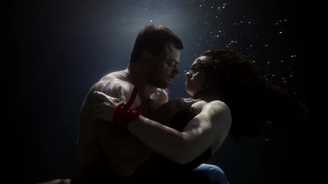 A sensual couple swim and spin under the dark water. A man gently and passionately embraces a woman in a black suit. There are a lot of bubbles floating around.