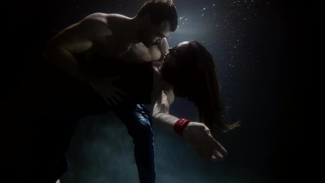 Sensual couple swim underwater. She has a collar around her neck and a bracelet on her arms. A man passionately embraces a woman. There are a lot of bubbles floating around.
