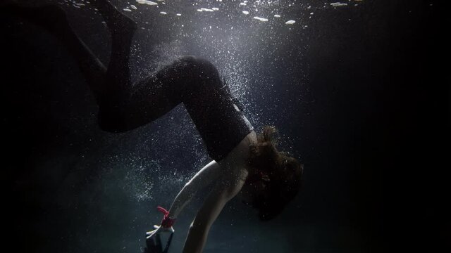 A woman in a suit is swimming underwater, doing a somersault in the water, her long hair flying. A mermaid and a nymph. She has a collar around her neck. There are a lot of bubbles floating around.