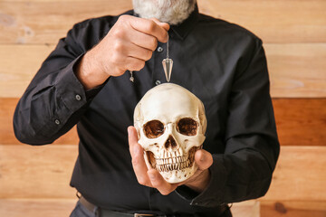 Male fortune teller with human skull and pendulum on wooden background