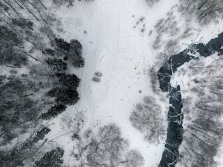 A snow-covered meadow with parked cars at the edge of a coniferous forest by the river. Aerial top down view of the nature cold winter landscape with two vehicle.