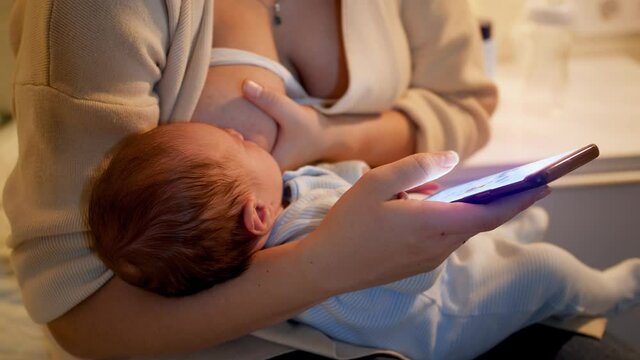 Closeup of mother feeding her little baby boy with breast milk holding smartphone and browsing social media. Concept of healthy and natural baby nutrition. Health of mother and child. Parenting and