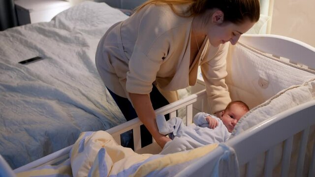 Beautiful young caring mother putting her newborn baby boy in crib and covering him with warm blanket at night. Concept of happy parenting and family happiness