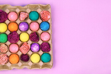 Multicolored Easter eggs painted with gouache and flowers in egg box on violet pink purple background, top view copy space