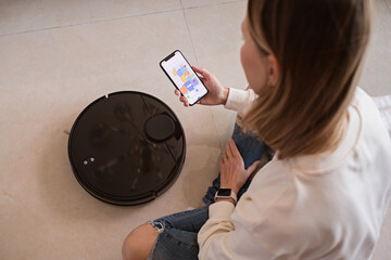 Robotic vacuum cleaner cleaning the room while woman relaxing. Woman controlling vacuum with remote...