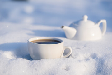 White cup of hot tea and teapot on a bed of snow and white background, close up
