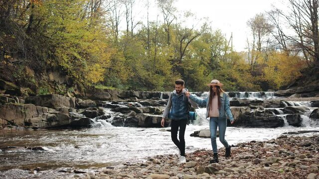 A young woman and a man are walking along the rocky shore of a mountain river. They are smiling. Hiking in the mountains. 4K.