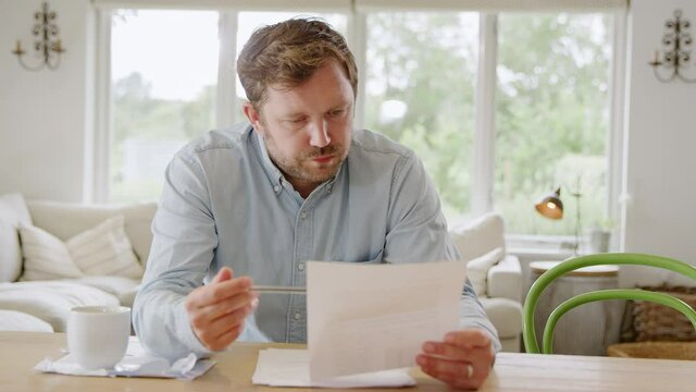 Worried man sitting at table at home reviewing domestic finances looking at bill - shot in slow motion