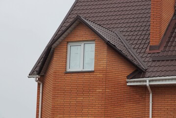 attic of a red brick private house with a white window under a brown tiled roof against a gray sky