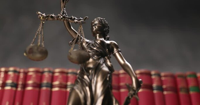 bronze law statue represented by blindfolded goddess spinning and holding a balance signifying impartiality and two broad sources of law with a background of books 