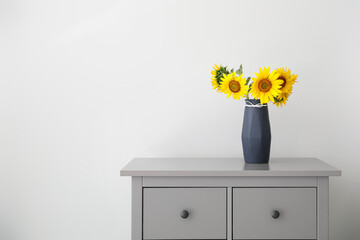 Bouquet of beautiful sunflowers in vase on commode near light wall. Space for text