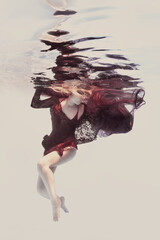 A woman in a red dress with ribbons and airy fabric poses underwater against a light background as if in zero gravity.