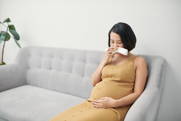 Pregnancy and illness. Sick pregnant woman blowing nose in tissue having fever sitting on sofa...